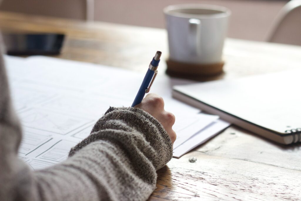 étudiant qui écrit avec crayon stylo sur des feuilles blanches en désordre sur une table en bois avec une tasse blanche en face de lui et un carnet beaucoup de luminosité