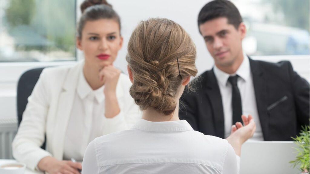 étudiante qui passe un oral d'école de commerce à Nantes en chemise blanche avec les cheveux attachés en chignon en face d'un jury composé d'un homme aux cheveux noir habillé en costume et une femme en costume blanc avec un rouge à lèvres rouge