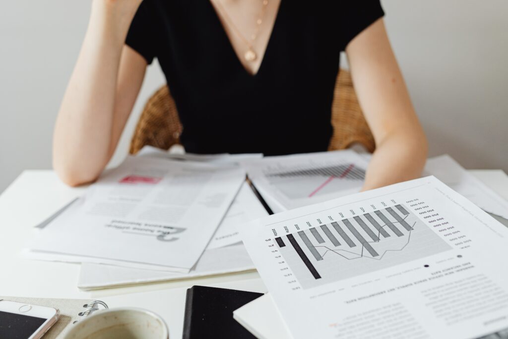 femme tee-shirt noir qui corrige des mémoires, plein de feuilles blanches en face d'elle sur une table, sur les feuilles il y a des écritures, des paragraphes et des graphiques