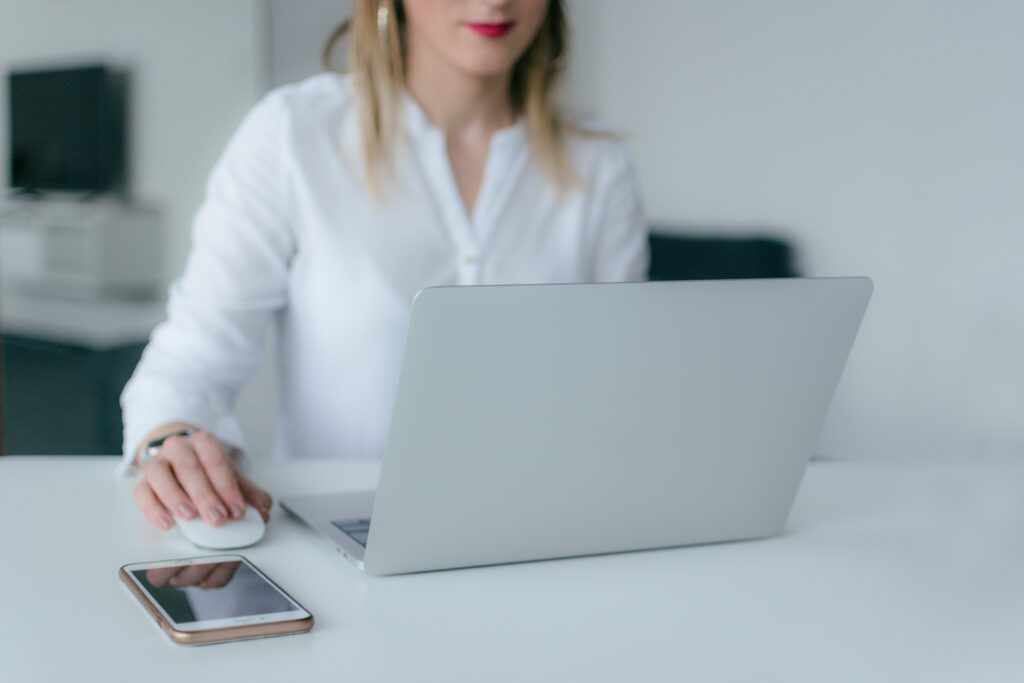 étudiante avec un haut blanc et du rouge à lèvres qui regarde son ordinateur avec une souris et un téléphone posé sur la table. 