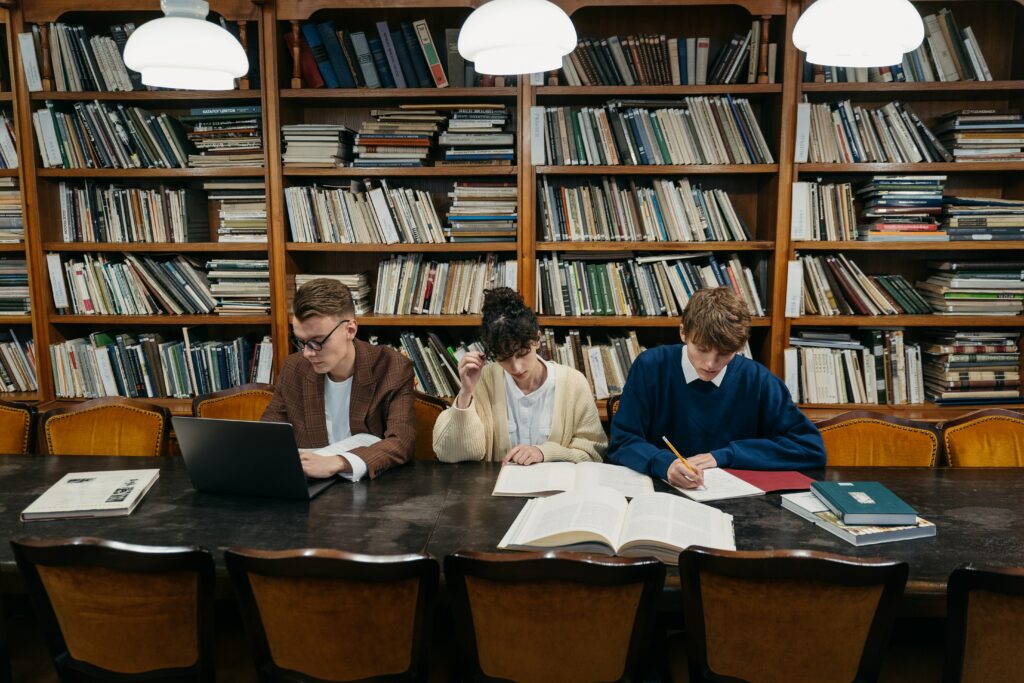 étudiants à la bibliothèque avec un garçon qui écrit au crayon sur un livre, une fille avec un gilet beige qui lit un livre et un autre étudiant avec des lunettes sur un ordinateur