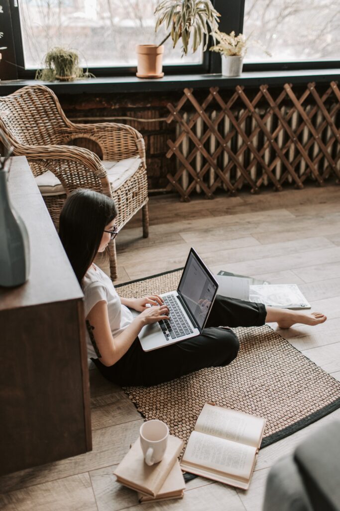 Femme qui écrit sur son ordinateur assise par terre sur un tapis marron avec un livre ouvert au sol et une tasse à côté 